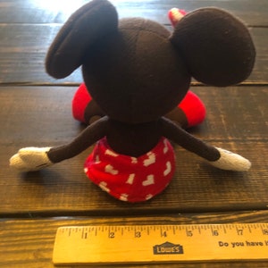 K&ouml;nnte beinhalten: Ein braunes und rotes Pl&uuml;sch-Mickey-Mouse-Spielzeug mit wei&szlig;en Handschuhen. Das Spielzeug sitzt auf einer Holzoberfl&auml;che.