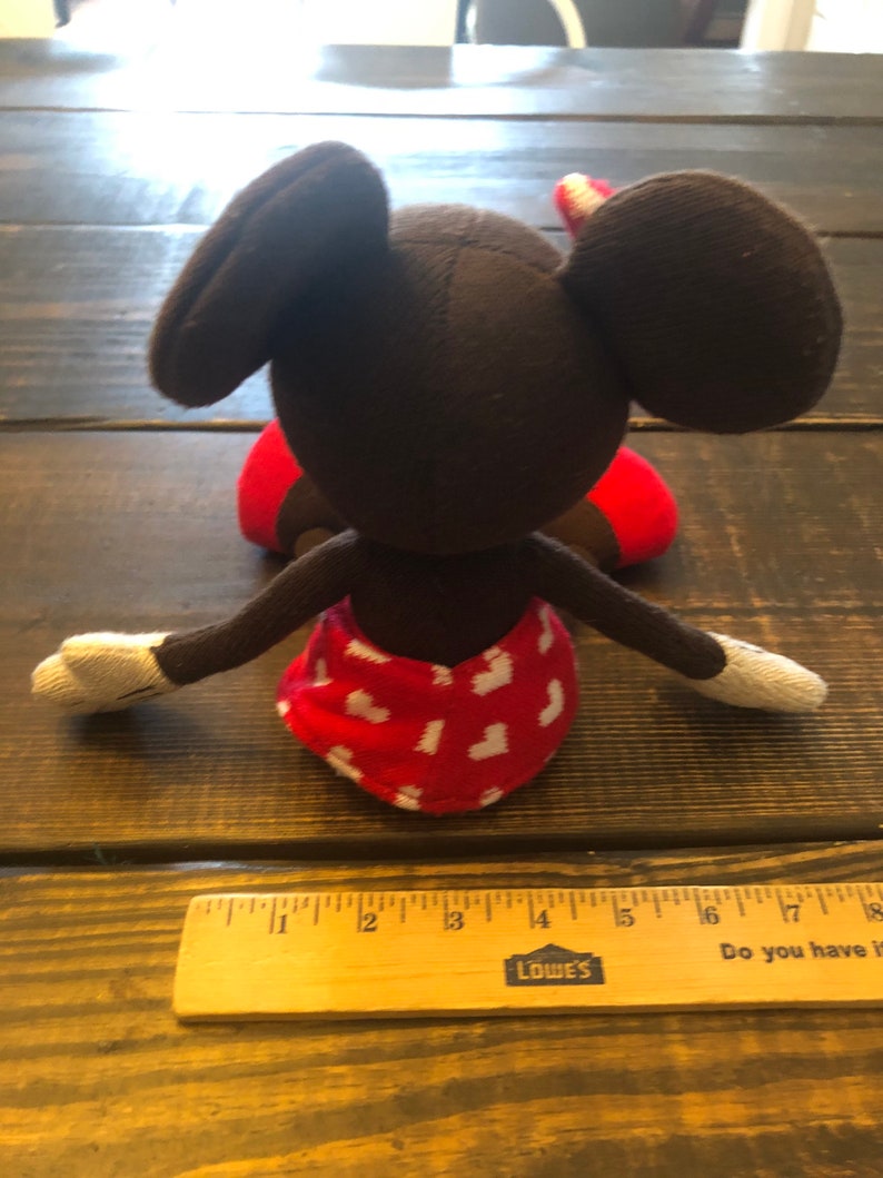 K&ouml;nnte beinhalten: Ein braunes und rotes Pl&uuml;sch-Mickey-Mouse-Spielzeug mit wei&szlig;en Handschuhen. Das Spielzeug sitzt auf einer Holzoberfl&auml;che.