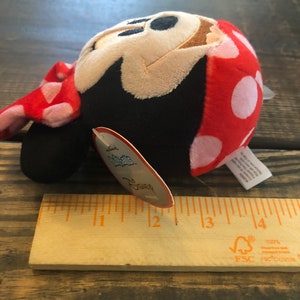 Op de afbeelding: Een pluche Minnie Mouse speelgoed in rood en wit met witte stippen, een zwart gezicht en een bruine neus. Het speelgoed is ongeveer 7,5 cm hoog en heeft een label met de tekst "Disney".