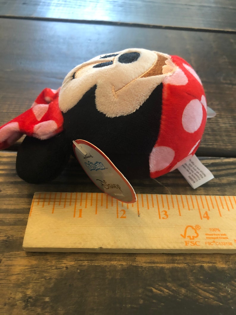 Op de afbeelding: Een pluche Minnie Mouse speelgoed in rood en wit met witte stippen, een zwart gezicht en een bruine neus. Het speelgoed is ongeveer 7,5 cm hoog en heeft een label met de tekst "Disney".