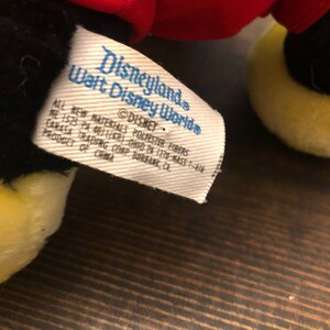 Puede incluir: Una etiqueta blanca con texto negro que dice "Disneyland Walt Disney World" y "DISNEY" con informaci&oacute;n adicional sobre los materiales y el origen del producto. La etiqueta est&aacute; unida a un juguete de peluche negro y amarillo.