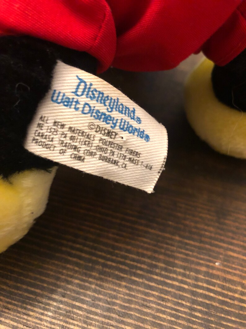 Puede incluir: Una etiqueta blanca con texto negro que dice "Disneyland Walt Disney World" y "DISNEY" con informaci&oacute;n adicional sobre los materiales y el origen del producto. La etiqueta est&aacute; unida a un juguete de peluche negro y amarillo.