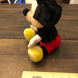 Puede incluir: Un juguete de peluche de Mickey Mouse sentado en una superficie de madera. El juguete es negro con pantalones cortos rojos y zapatos amarillos. El juguete mide aproximadamente 15 cm de alto.