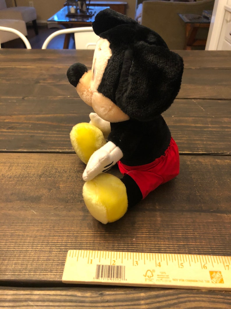 Puede incluir: Un juguete de peluche de Mickey Mouse sentado en una superficie de madera. El juguete es negro con pantalones cortos rojos y zapatos amarillos. El juguete mide aproximadamente 15 cm de alto.