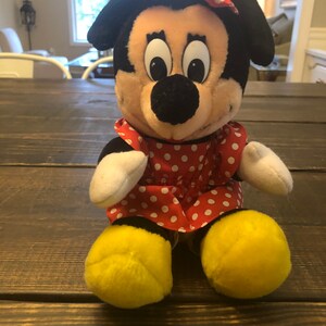 May include: A plush toy of Minnie Mouse wearing a red dress with white polka dots and a red bow with white polka dots. She has yellow shoes and a black nose.