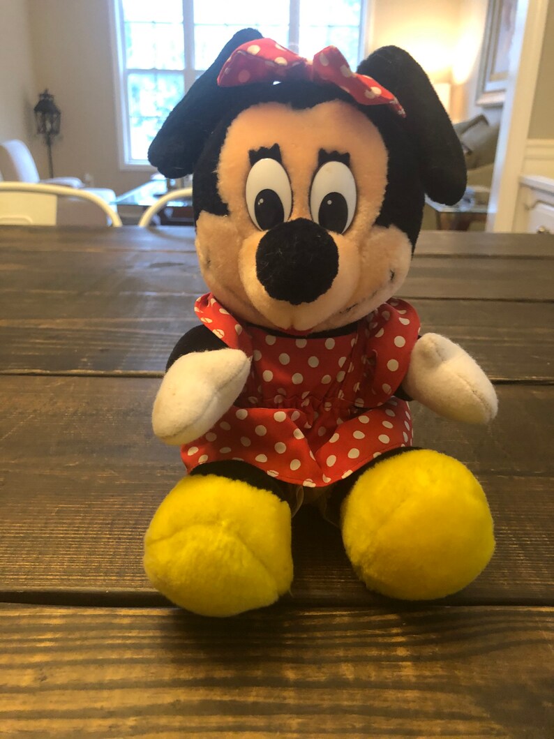May include: A plush toy of Minnie Mouse wearing a red dress with white polka dots and a red bow with white polka dots. She has yellow shoes and a black nose.