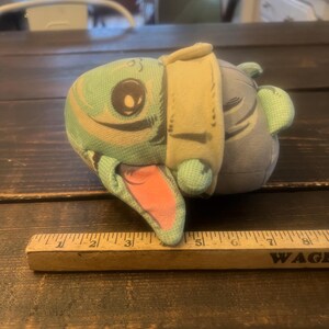 Pode incluir: Um brinquedo de pel&uacute;cia em forma de personagem de desenho animado, com um rosto verde claro, um olho grande e uma boca rosa. O brinquedo tem uma roupa bege e um corpo cinza. Uma r&eacute;gua de madeira &eacute; colocada perto.