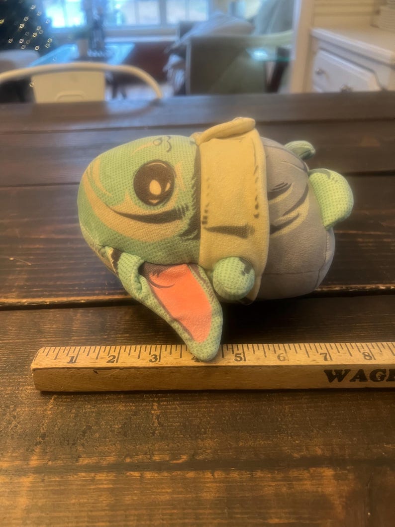 Pode incluir: Um brinquedo de pel&uacute;cia em forma de personagem de desenho animado, com um rosto verde claro, um olho grande e uma boca rosa. O brinquedo tem uma roupa bege e um corpo cinza. Uma r&eacute;gua de madeira &eacute; colocada perto.