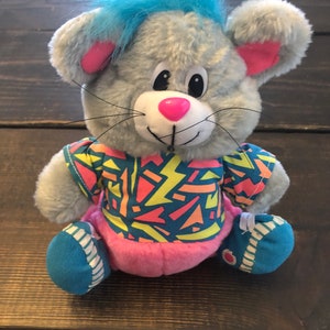 May include: A gray plush mouse with blue hair, wearing a pink, yellow, and blue shirt with a geometric pattern. The mouse is wearing blue and white sneakers.