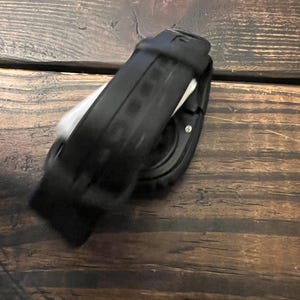 May include: Black smartwatch with a black band. The watch is lying on a wooden surface. The watch band is made of a flexible material and has a textured design. The watch face is round and has a dark display.