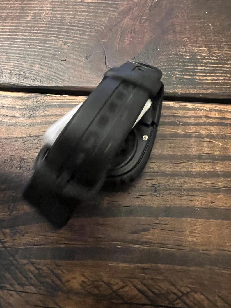 May include: Black smartwatch with a black band. The watch is lying on a wooden surface. The watch band is made of a flexible material and has a textured design. The watch face is round and has a dark display.