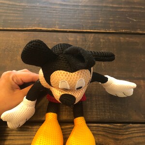 May include: A black and yellow mesh Mickey Mouse plush toy. The toy has a red shirt and large black ears.