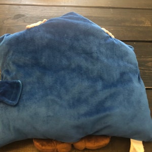 以下が含まれることがあります： 青いぬいぐるみ枕。枕の下から茶色のキャラクターが顔を出しています。キャラクターは茶色の足が2本と手が2本あります。枕は柔らかくふわふわした素材でできています。