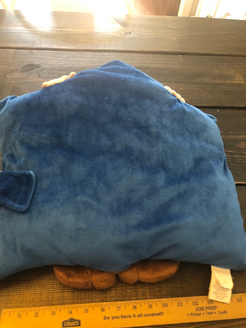 以下が含まれることがあります： 青いぬいぐるみ枕。枕の下から茶色のキャラクターが顔を出しています。キャラクターは茶色の足が2本と手が2本あります。枕は柔らかくふわふわした素材でできています。