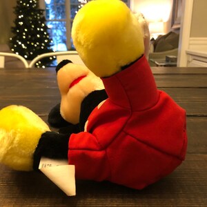 May include: A red and yellow plush Mickey Mouse toy lying on a wooden surface. The toy is wearing a red jacket with black buttons and has a yellow face with black eyes and a red nose.