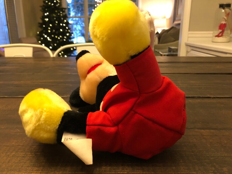 May include: A red and yellow plush Mickey Mouse toy lying on a wooden surface. The toy is wearing a red jacket with black buttons and has a yellow face with black eyes and a red nose.