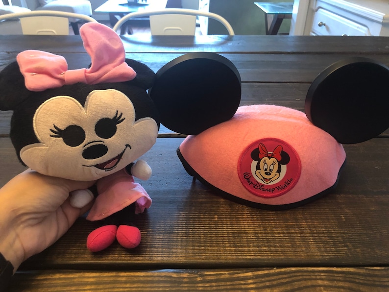 May include: Pink Minnie Mouse plush toy with a black bow and a pink dress. Next to it is a pink Minnie Mouse ear headband with black ears and a black and white Walt Disney World logo.