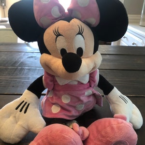 May include: A pink and black plush Minnie Mouse toy with white polka dots. She is wearing a pink dress with white polka dots and a pink bow. The toy has a Disney logo on the bottom of one foot.