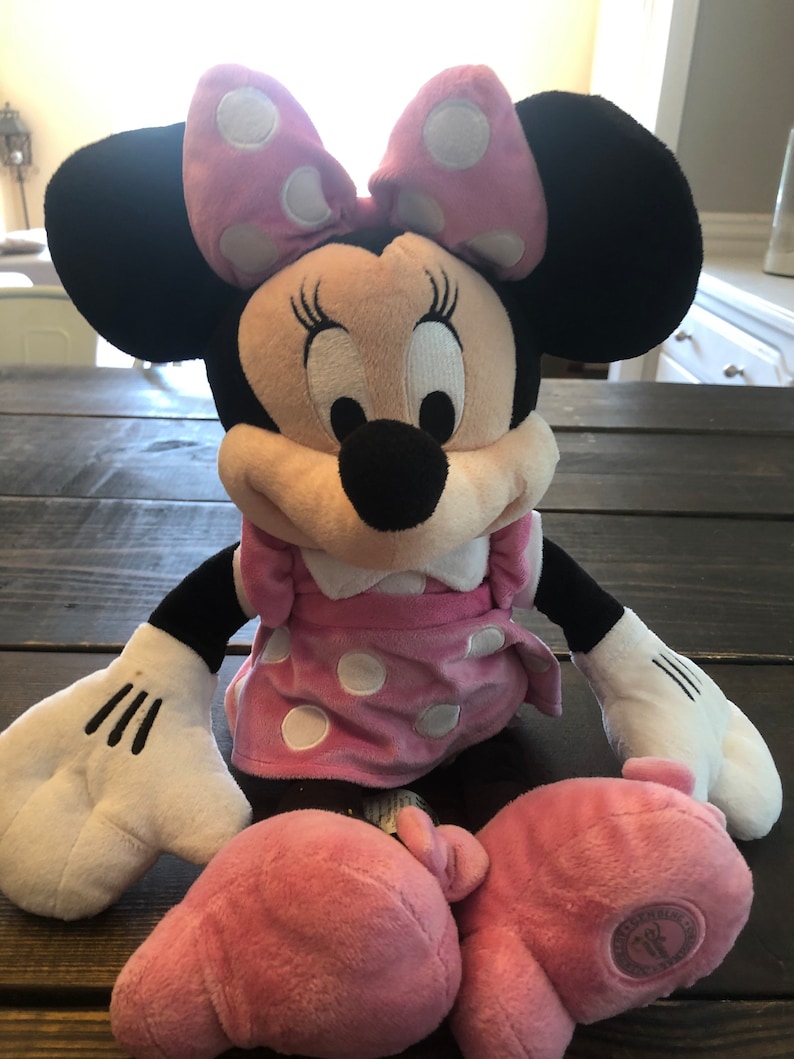 May include: A pink and black plush Minnie Mouse toy with white polka dots. She is wearing a pink dress with white polka dots and a pink bow. The toy has a Disney logo on the bottom of one foot.