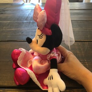 May include: Pink Minnie Mouse plush toy wearing a pink dress with white lace trim and a pink hat with a white veil. The toy is sitting on a wooden surface.