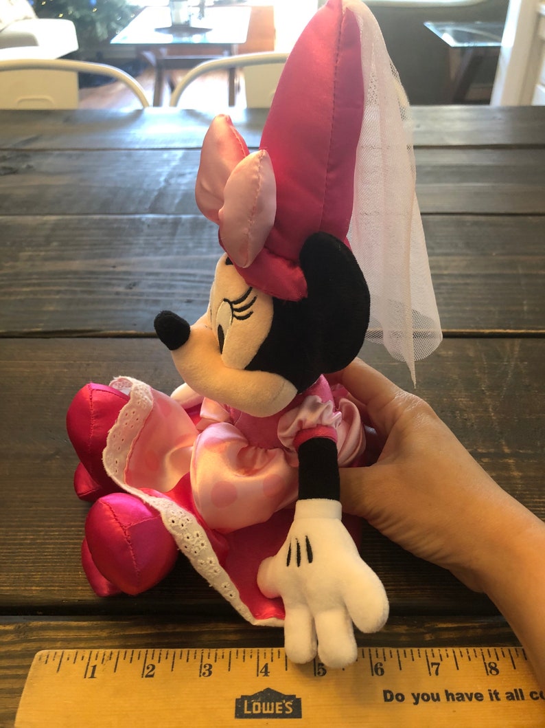 May include: Pink Minnie Mouse plush toy wearing a pink dress with white lace trim and a pink hat with a white veil. The toy is sitting on a wooden surface.