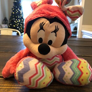 Op de afbeelding: Een roze pluche speelgoed van Minnie Mouse met een paasoutfit met een roze hoed en een kleurrijke chevron-broek. Het speelgoed heeft een zwarte neus en grote ogen.
