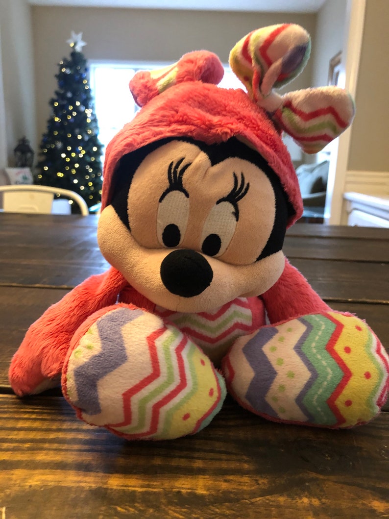 Op de afbeelding: Een roze pluche speelgoed van Minnie Mouse met een paasoutfit met een roze hoed en een kleurrijke chevron-broek. Het speelgoed heeft een zwarte neus en grote ogen.