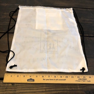 May include: White drawstring bag with black drawstrings and a white logo that says "Remarkable Arts" printed on the front.