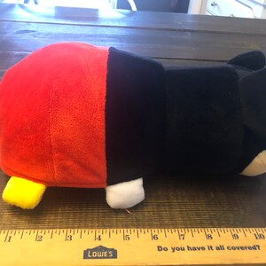 May include: A red and black plush toy shaped like Mickey Mouse. The toy is lying on its side with its head turned to the right. The toy has a yellow nose and white eyes.