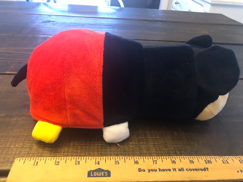 May include: A red and black plush toy shaped like Mickey Mouse. The toy is lying on its side with its head turned to the right. The toy has a yellow nose and white eyes.