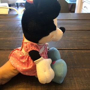 May include: A plush Minnie Mouse toy wearing a pink polka dot dress and blue shoes. The toy is sitting on a wooden surface with a ruler in the foreground.