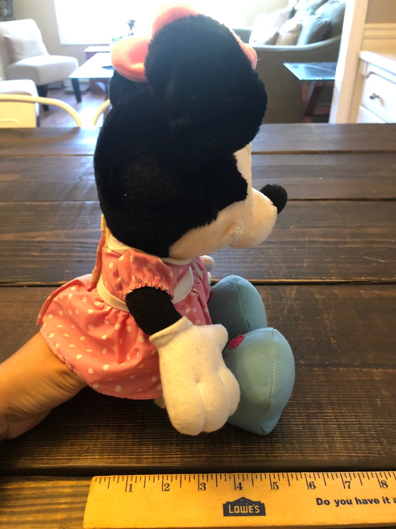 May include: A plush Minnie Mouse toy wearing a pink polka dot dress and blue shoes. The toy is sitting on a wooden surface with a ruler in the foreground.