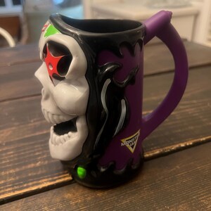 May include: A ceramic mug featuring a skull design with a purple handle. The mug has a white skull face with red star accents, green and black details, and the Monster Jam logo. The mug is designed with a purple background.