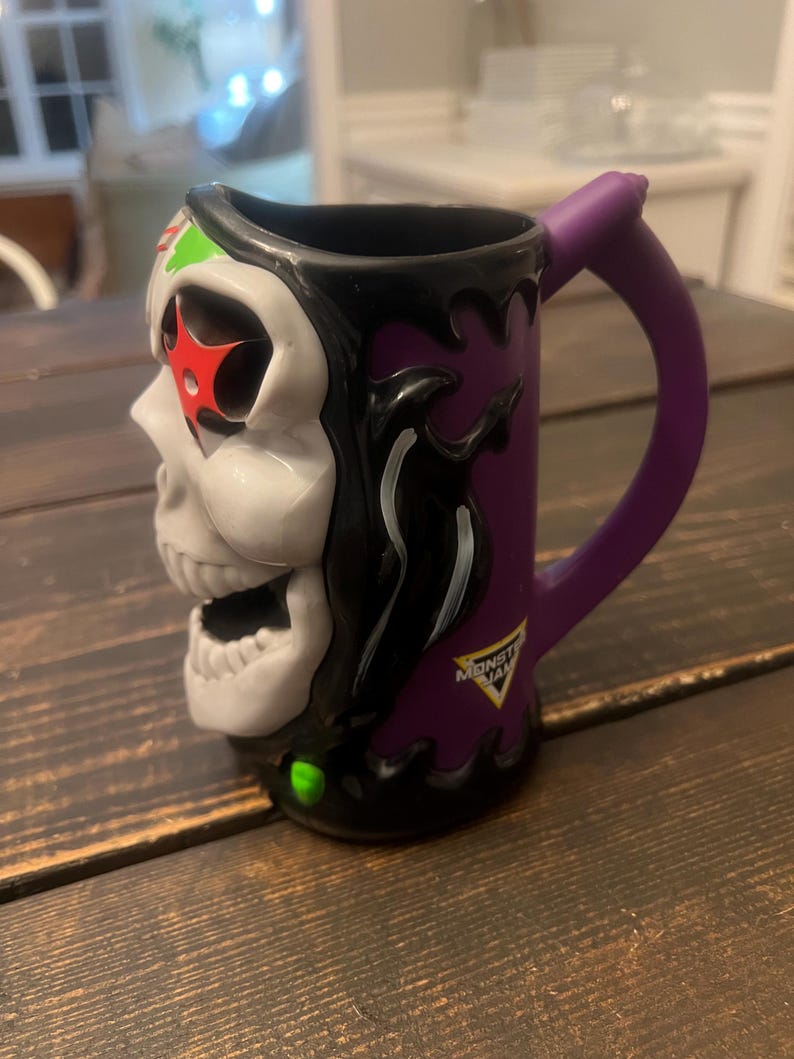 May include: A ceramic mug featuring a skull design with a purple handle. The mug has a white skull face with red star accents, green and black details, and the Monster Jam logo. The mug is designed with a purple background.