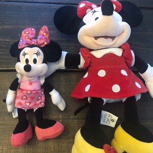 May include: Two plush Minnie Mouse toys. The larger Minnie Mouse is wearing a red dress with white polka dots and yellow shoes with red bows. The smaller Minnie Mouse is wearing a pink dress with red hearts and pink shoes. Both Minnie Mouse toys have black ears and red bows.