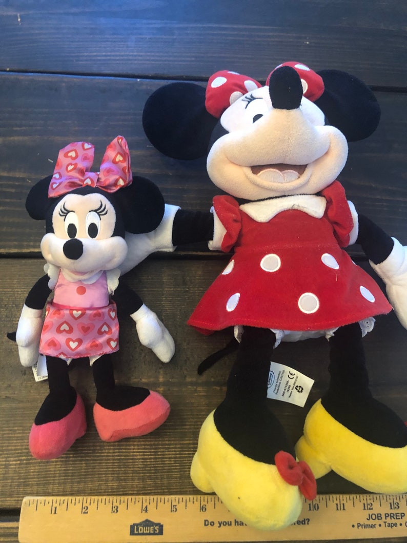 May include: Two plush Minnie Mouse toys. The larger Minnie Mouse is wearing a red dress with white polka dots and yellow shoes with red bows. The smaller Minnie Mouse is wearing a pink dress with red hearts and pink shoes. Both Minnie Mouse toys have black ears and red bows.