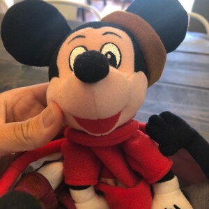 Peut inclure: Un jouet en peluche Mickey Mouse portant une robe rouge et un chapeau marron. Le jouet a un grand sourire et de grands yeux noirs.