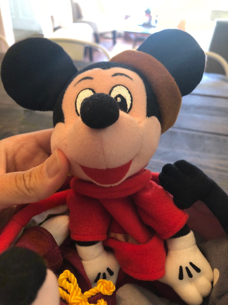 Peut inclure: Un jouet en peluche Mickey Mouse portant une robe rouge et un chapeau marron. Le jouet a un grand sourire et de grands yeux noirs.