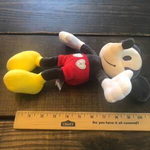 Puede incluir: Un juguete de peluche de Mickey Mouse con una camisa roja y zapatos amarillos. El juguete est&aacute; acostado sobre una superficie de madera con una regla a su lado. La regla tiene las palabras "LOWE'S" y "Do you have it all covered?" impresas en ella.