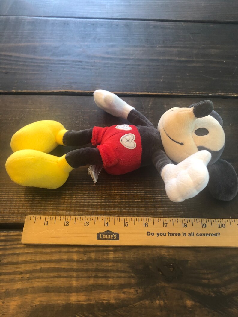 Puede incluir: Un juguete de peluche de Mickey Mouse con una camisa roja y zapatos amarillos. El juguete est&aacute; acostado sobre una superficie de madera con una regla a su lado. La regla tiene las palabras "LOWE'S" y "Do you have it all covered?" impresas en ella.