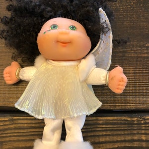 May include: A Cabbage Patch Kid doll dressed as an angel. The doll has black curly hair, green eyes, and a white dress with a gold glitter overlay. The doll is wearing white pants and white boots with gold accents.