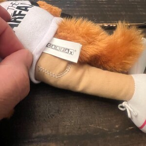 May include: A plush toy with orange fur, beige trousers, and white shoes. The toy is wearing a white shirt with the text "THE GREAT" on it. A tag with the "CARFAX" logo is attached to the toy.