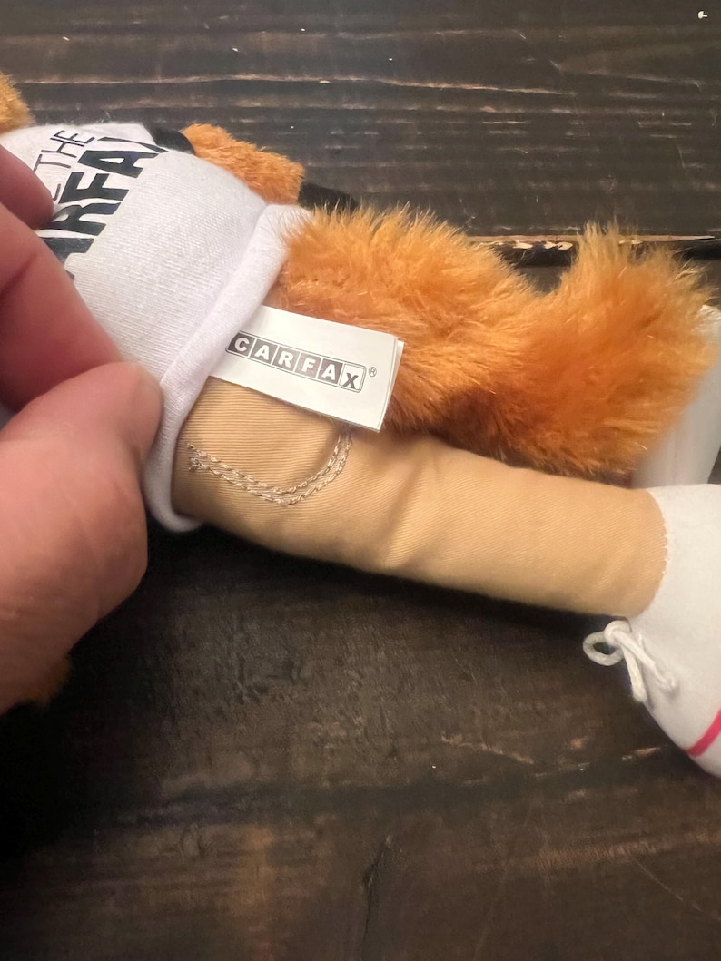 May include: A plush toy with orange fur, beige trousers, and white shoes. The toy is wearing a white shirt with the text "THE GREAT" on it. A tag with the "CARFAX" logo is attached to the toy.