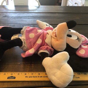 May include: Pink and black plush Minnie Mouse toy with white polka dots on her dress and bow. She is lying on a wooden surface with a ruler in the foreground.