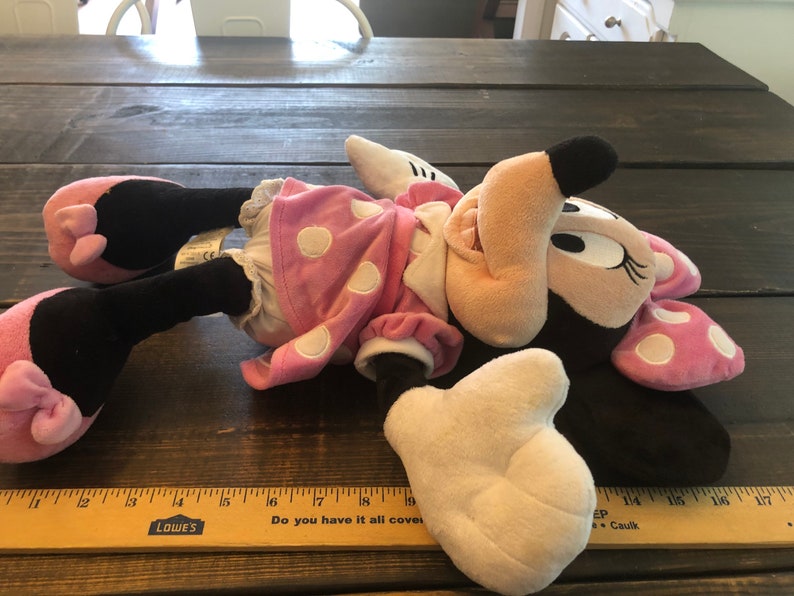 May include: Pink and black plush Minnie Mouse toy with white polka dots on her dress and bow. She is lying on a wooden surface with a ruler in the foreground.