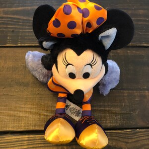 May include: A Minnie Mouse plush toy with a black head and ears, a light pink face, and a black nose. She wears an orange bow with purple polka dots, an orange and purple striped outfit, and gold shoes. The toy has a Disney tag.
