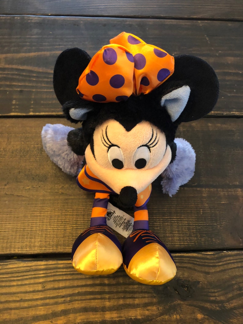 May include: A Minnie Mouse plush toy with a black head and ears, a light pink face, and a black nose. She wears an orange bow with purple polka dots, an orange and purple striped outfit, and gold shoes. The toy has a Disney tag.