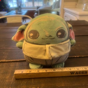 Pode incluir: Um brinquedo de pel&uacute;cia de um personagem com olhos grandes, pele verde e orelhas grandes. O brinquedo est&aacute; vestindo uma roupa bege. Uma r&eacute;gua de madeira com a palavra "WAGES" est&aacute; em primeiro plano.