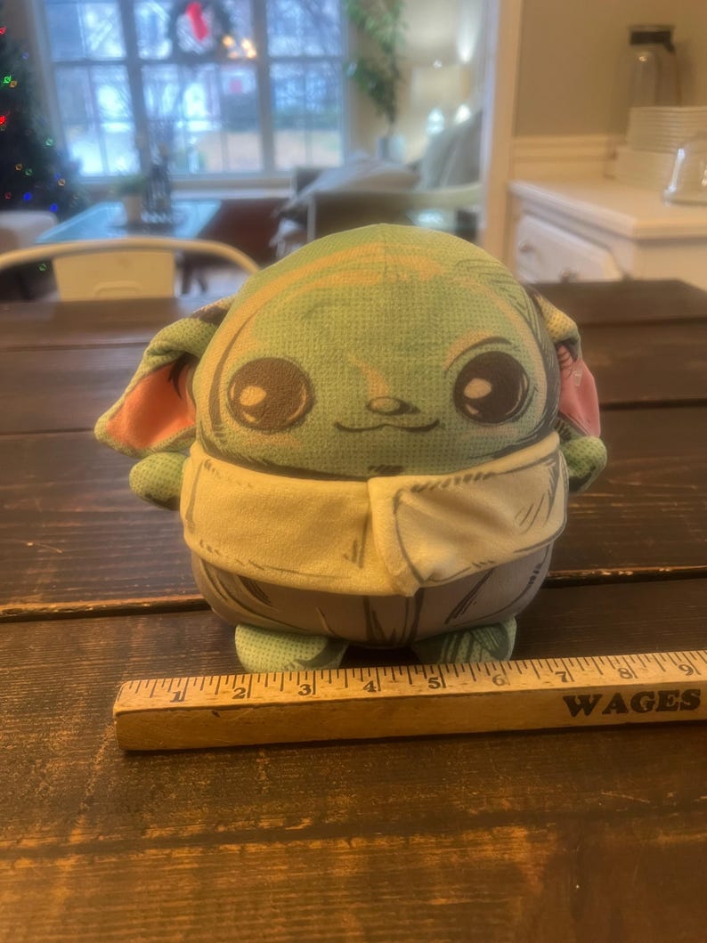 Pode incluir: Um brinquedo de pel&uacute;cia de um personagem com olhos grandes, pele verde e orelhas grandes. O brinquedo est&aacute; vestindo uma roupa bege. Uma r&eacute;gua de madeira com a palavra "WAGES" est&aacute; em primeiro plano.