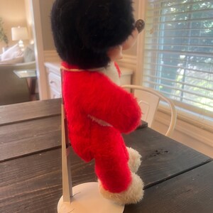 May include: A vintage plush toy of a mouse character, dressed in a red coat with white trim and a black furry hat. The toy is mounted on a white metal stand, showcasing its side profile. The toy's features include a round black nose and small ears.